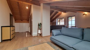 A spacious living area with a high ceiling, wooden beams, a blue sofa, and a kitchen in the background. - New Listing (Riegsee)