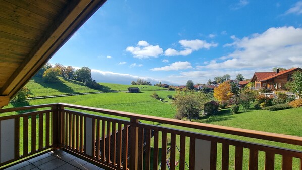 A wooden balcony with a view of a green landscape, houses, and a clear sky. - New Listing (Riegsee)