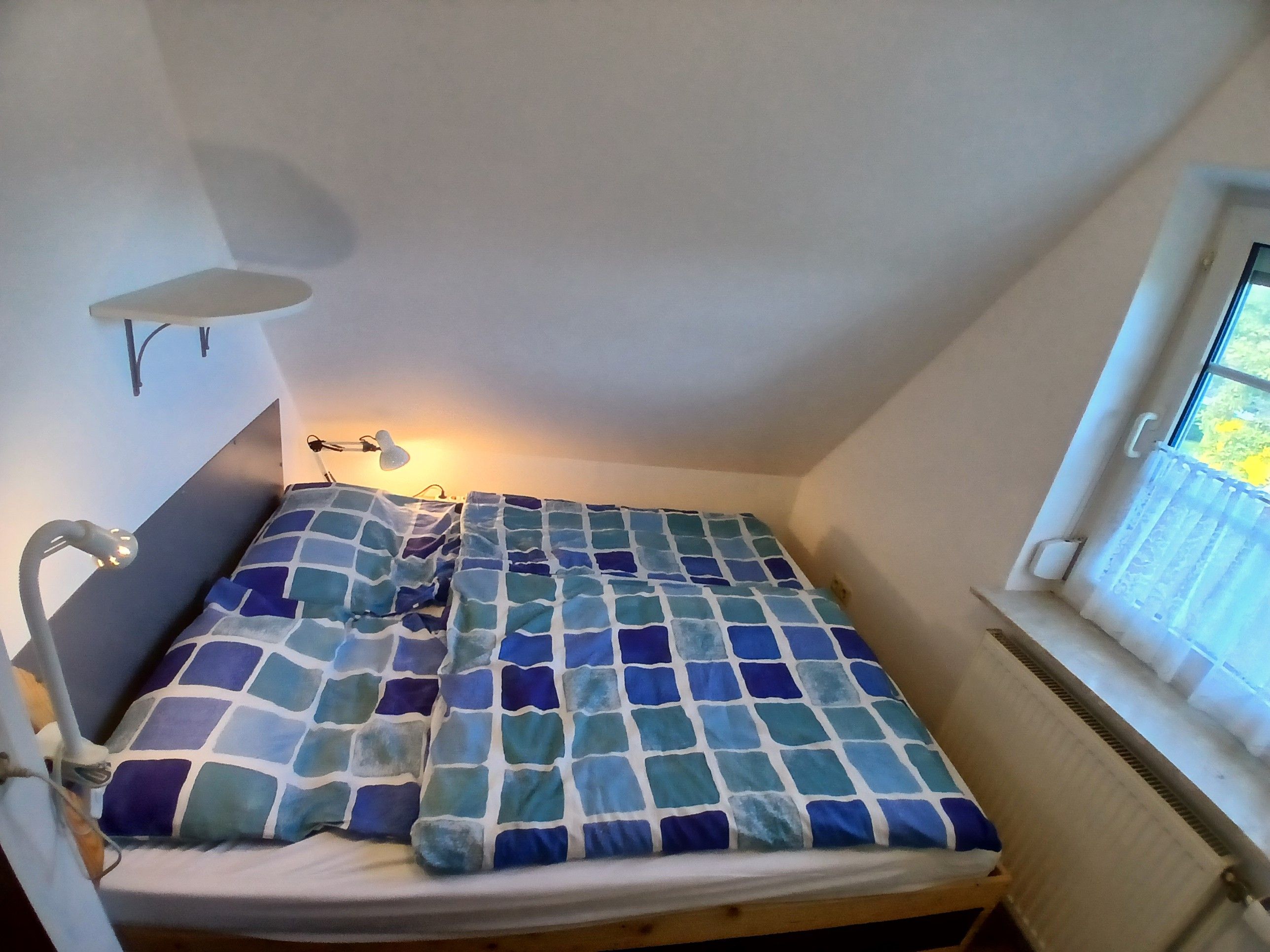 A small, single-bed room with a headboard, a bedside lamp, and a window with a view of greenery outside.