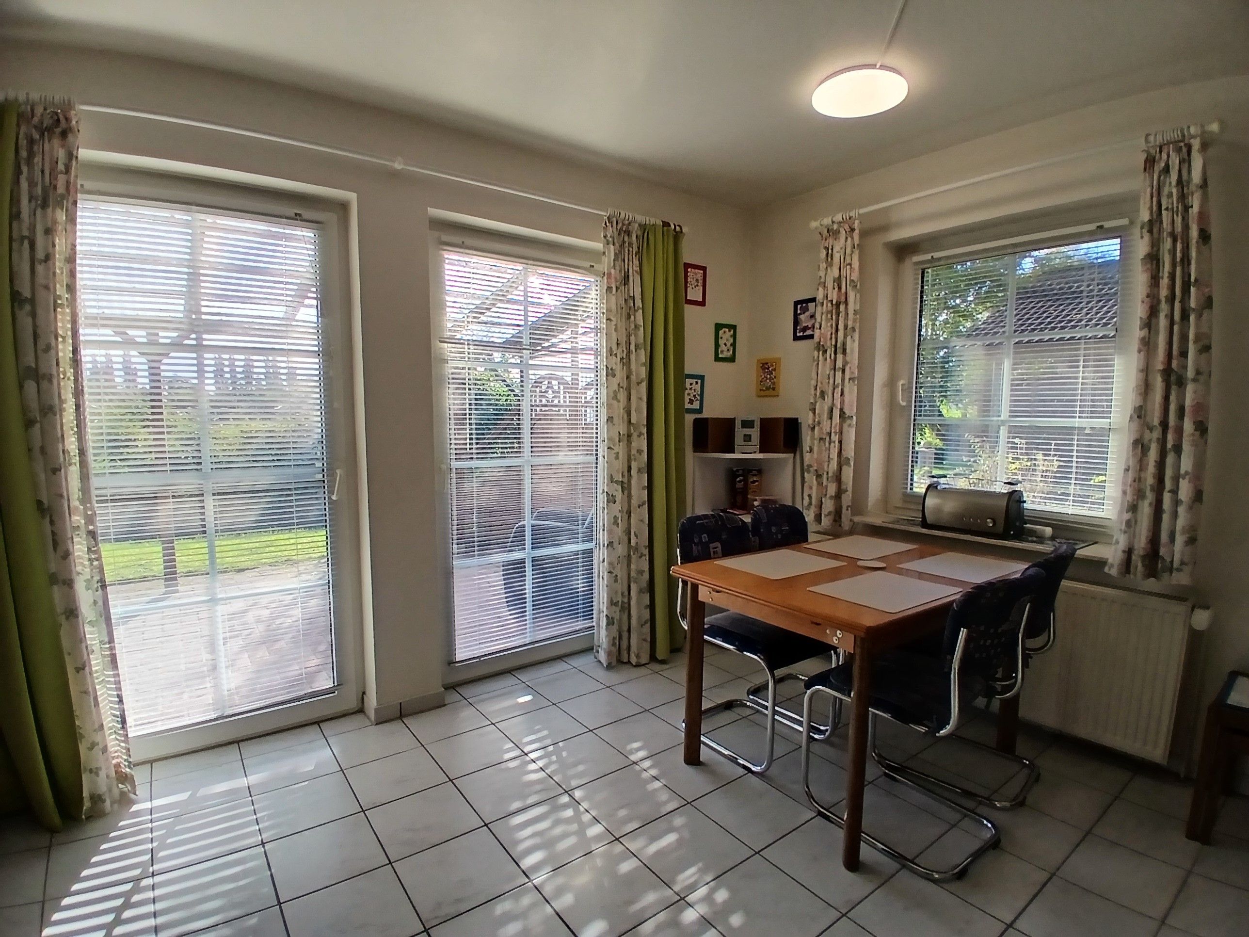 A room with a dining table, chairs, a radiator, and large windows with curtains.