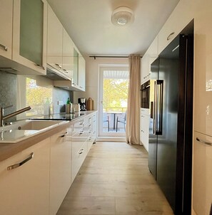 Eine schmale Küche mit Kühlschrank, Schränken und einem Fenster zum Balkon.