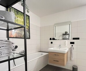 Ein modernes Badezimmer mit einer Badewanne, einem Waschbecken und einem Spiegel. Ein Regal mit Handtüchern und ein gerahmtes Bild sind ebenfalls vorhanden.