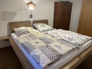 Un lit avec une couvre-lit matelassé et deux oreillers, une table de chevet, une lampe fixée au mur et une armoire en bois.