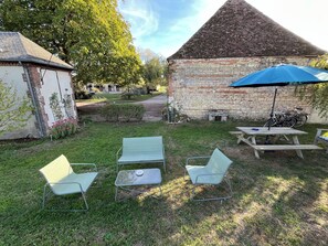 Eine grüne Rasenfläche mit Picknicktisch und -stühlen, einem blauen Sonnenschirm und einem Gebäude aus Stein mit Satteldach.