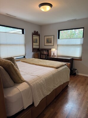 Quarto com cama, mesa de cabeceira, abajur, estante de livros e duas janelas com persianas.