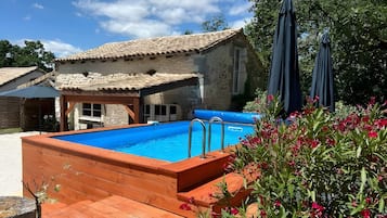 Une piscine avec une terrasse en bois, un bâtiment en pierre doté d’un toit en tuiles et un jardin parsemé de fleurs roses.