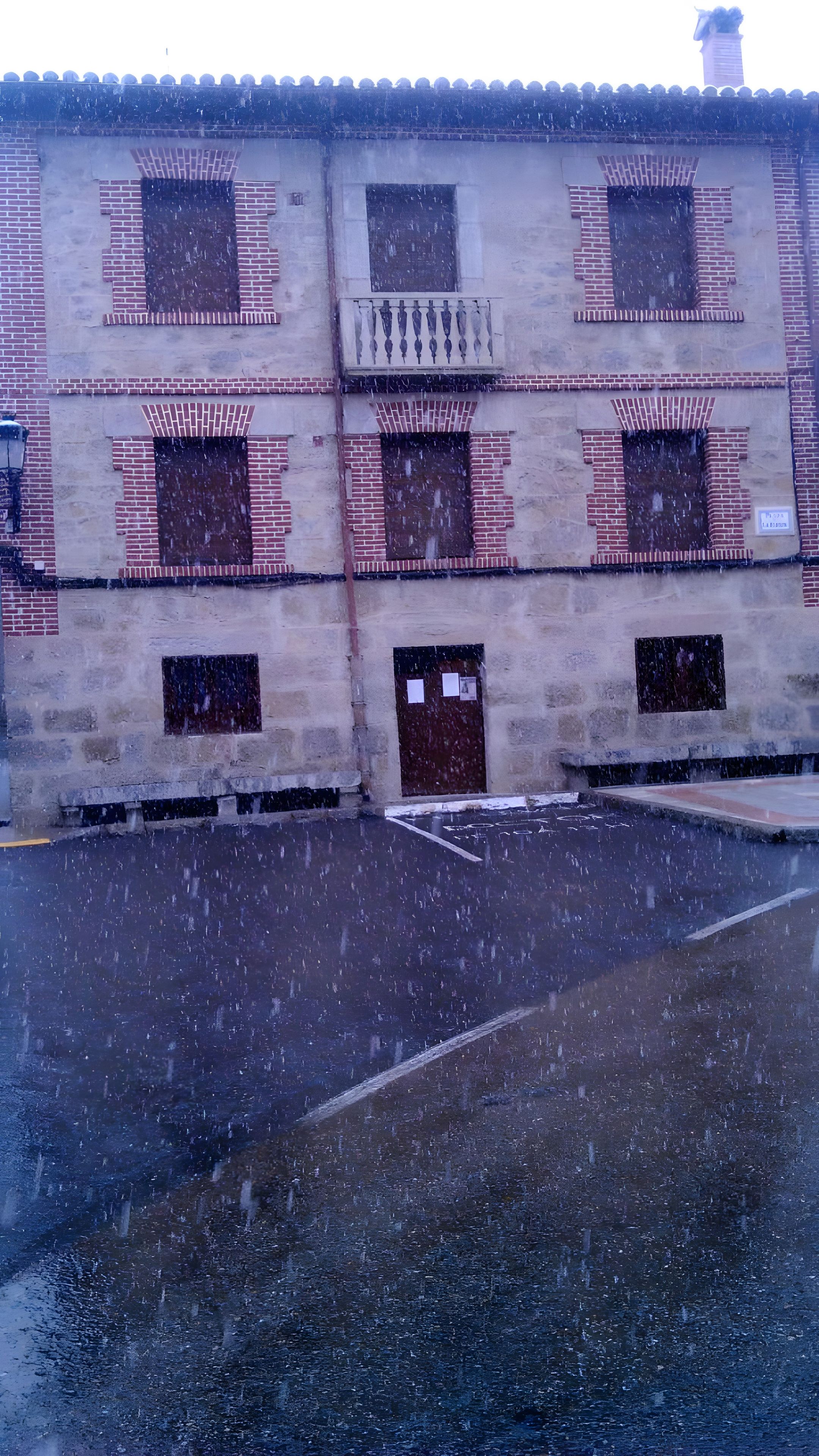 Un edificio con varias ventanas y puertas, algunas con contraventanas, y un área pavimentada en la parte delantera.