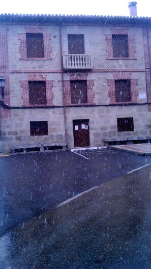 Un edificio con varias ventanas y puertas, algunas con contraventanas, y un área pavimentada en la parte delantera.