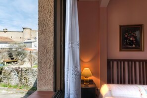 A hotel room with a bed, a nightstand with a lamp, a painting, and a view of a historic building. - Country House 'La Casa Grande' with Fireplace, Patio with Barbecue and Wi-Fi (El Barco de Ávila)
