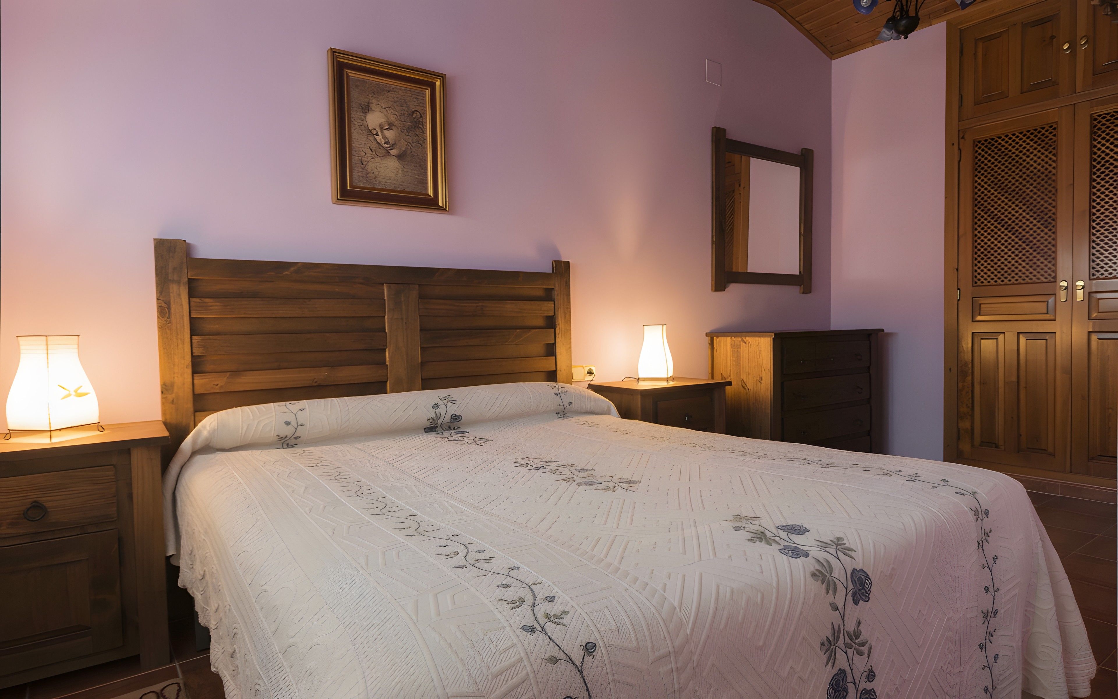A bedroom with a wooden headboard, a bed with floral bedding, bedside lamps, a wooden wardrobe, and a wooden door with a lattice design.