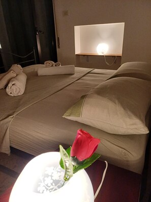 Ein Hotelzimmer mit einem ordentlich bezogenen Bett, einer Nachttischlampe und einer Rose auf dem Nachttisch.