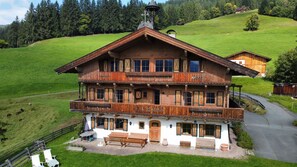 A two-story wooden chalet with a balcony, a paved patio, and a view of the surrounding green landscape. - New Listing (Kirchberg in Tirol)