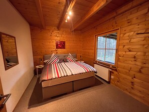 Ein gemütliches Schlafzimmer in einer Holzhütte mit Bett, Nachttisch, Spiegel und Fenster mit Blick auf Bäume.