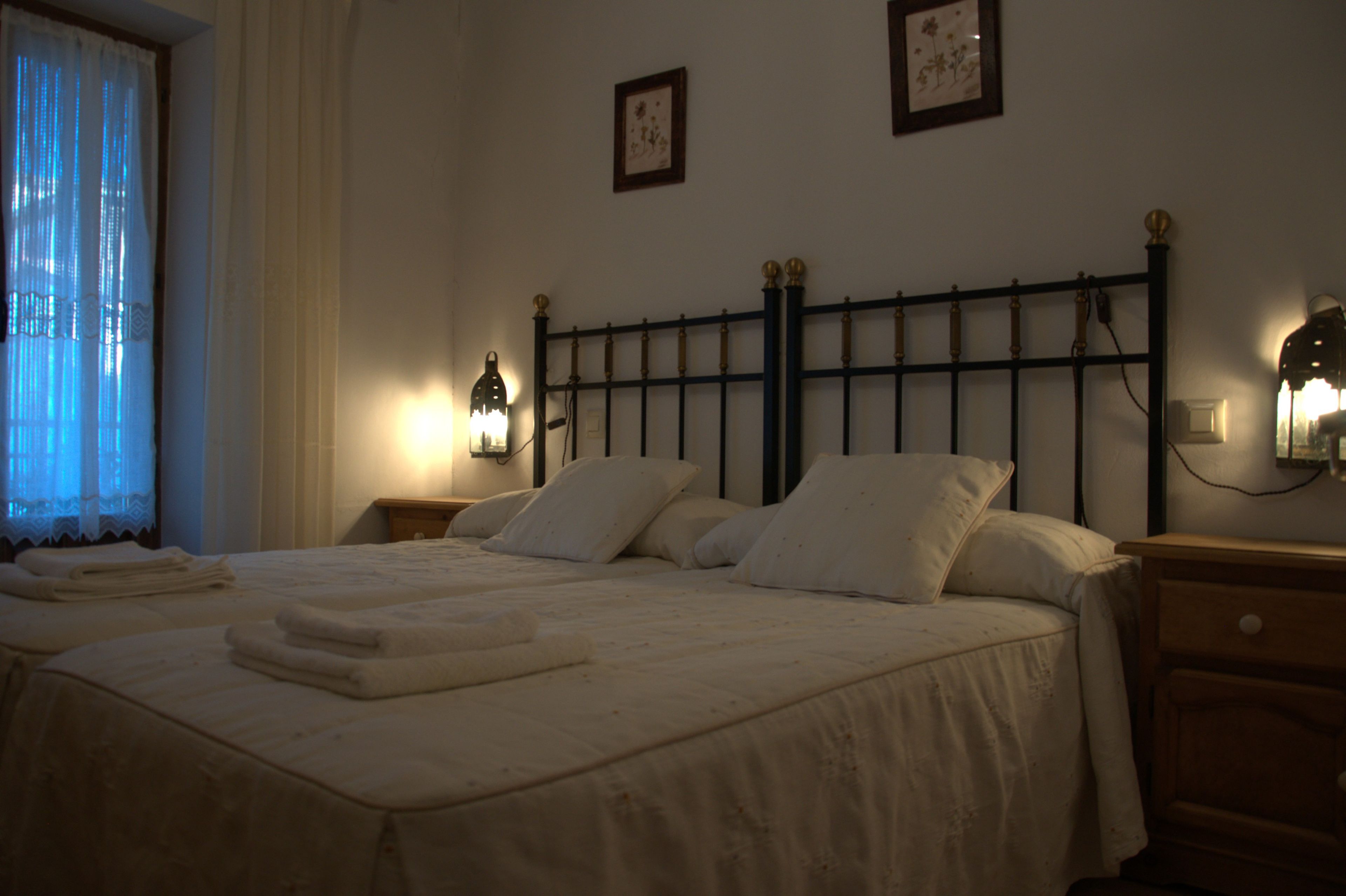 Habitación de hotel con una cama grande, dos mesitas de noche y un buró. Hay dos lámparas de pared y un ventanal con cortinas.