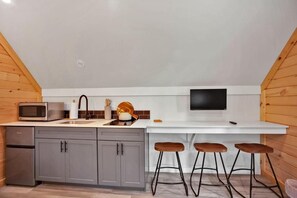 A compact kitchen with a microwave, sink, and a flat-screen TV. There are three stools at the counter. - PA Grand Canyon Cozy Cabin (Wellsboro)