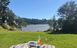A picnic setup with a table, a bottle, and a container on a grassy area with a lake and trees in the background. - A peaceful harbour-side retreat in the beautiful Eastern Bay of Plenty (Wainui)