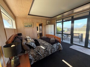 A bedroom with a bed, a nightstand, a lamp, a painting, and a view of the outdoors. - A peaceful harbour-side retreat in the beautiful Eastern Bay of Plenty (Wainui)