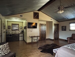 Um quarto espaçoso com piso de madeira, uma cama, um sofá de couro, uma televisão na parede e uma cozinha compacta com micro-ondas.