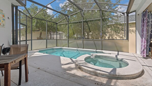 Una piscina con un jacuzzi, rodeada por un recinto de vidrio, con una silla y una mesa cerca.