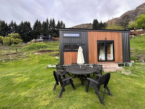 A modern cabin with a wooden exterior and a glass door, situated on a grassy hill with a view of a forested area.