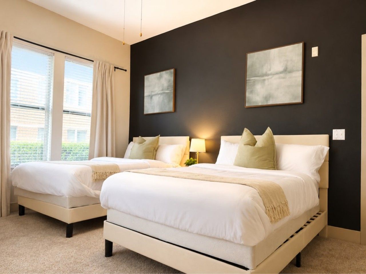 A hotel room with two beds, a large window with curtains, and two framed artworks on the wall.