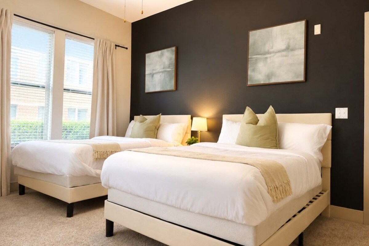 Een hotelkamer met twee bedden, een groot raam met gordijnen en twee ingelijste kunstwerken aan de muur.