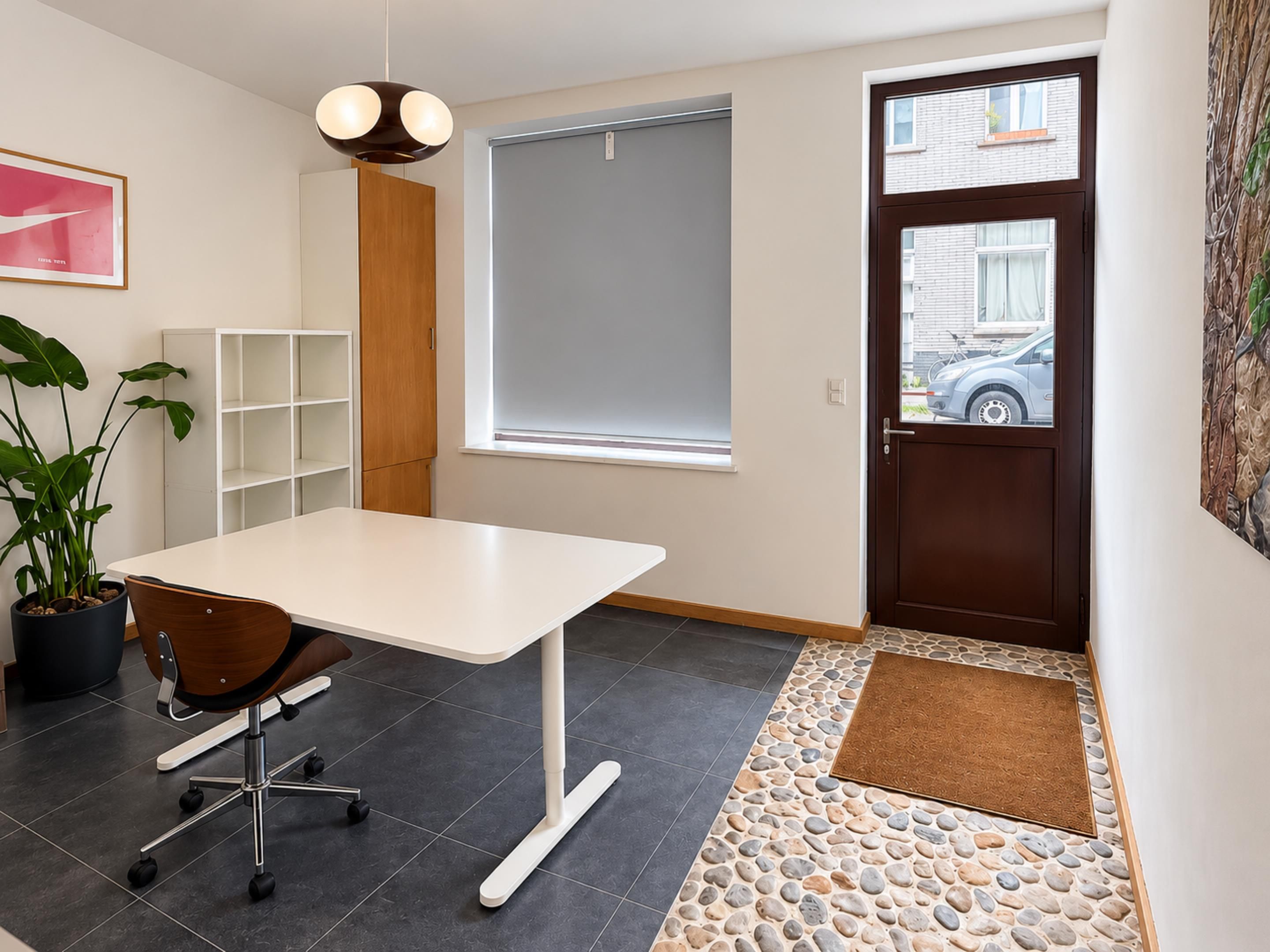 Un espace de bureau moderne avec un bureau blanc, une plante en pot, une bibliothèque, une fenêtre avec des stores et une porte donnant sur la rue.