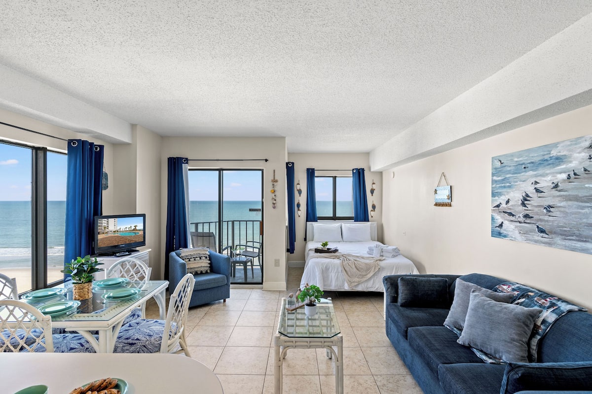 A spacious living area with a balcony overlooking the sea, a dining table with chairs, a sofa, and a television.
