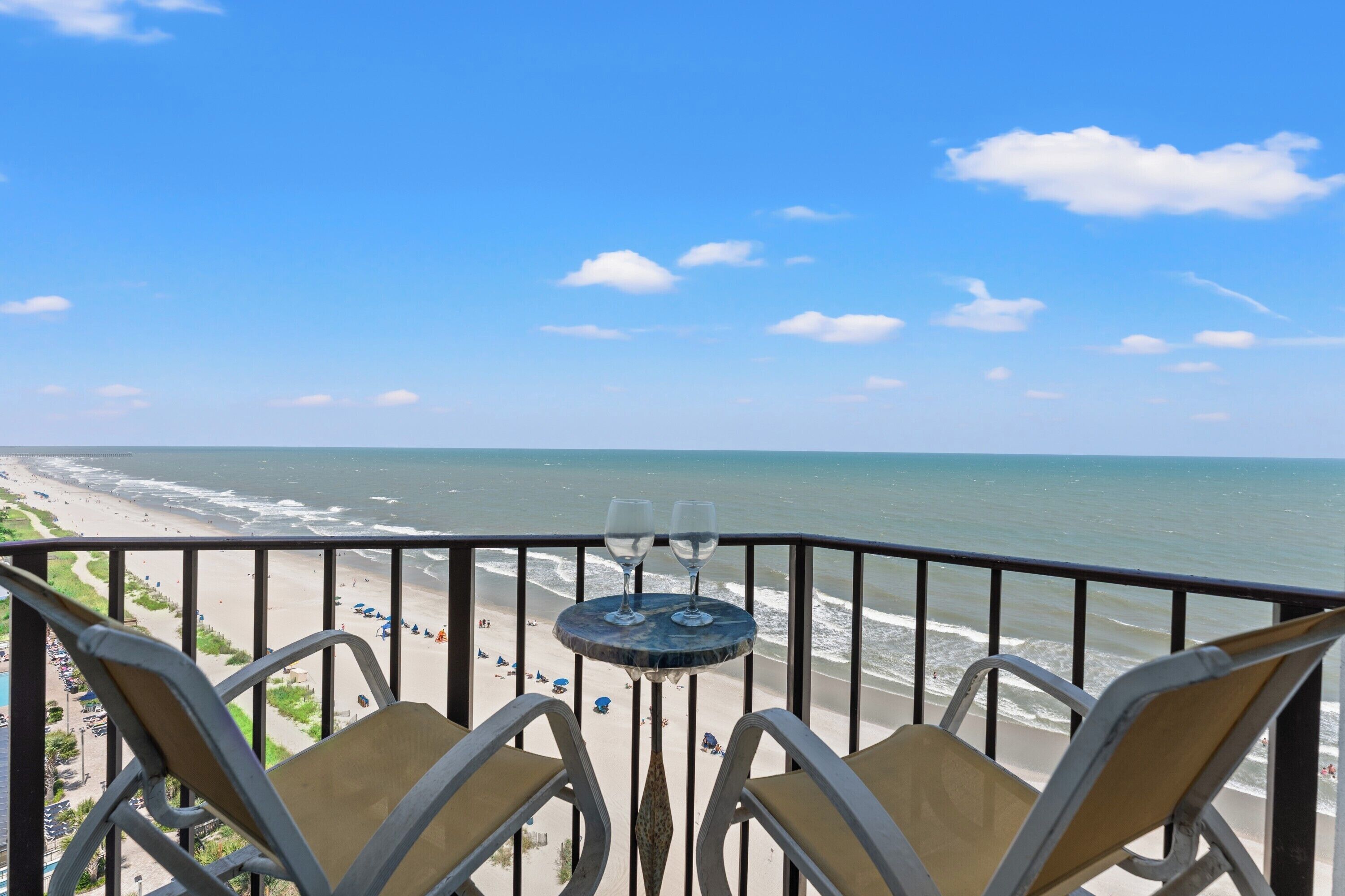 A balcony with two chairs and a table, overlooking a beach and ocean.