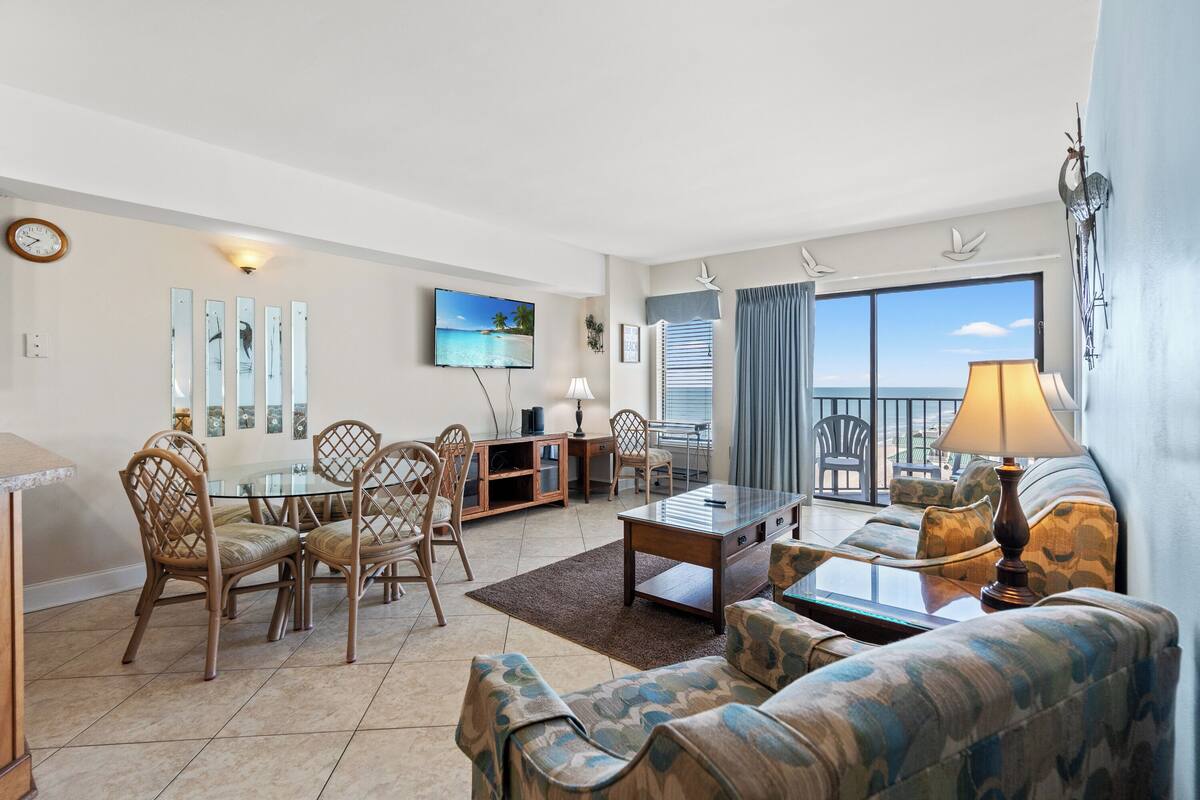 A spacious living room with a large TV, comfortable seating, and a view of the ocean.