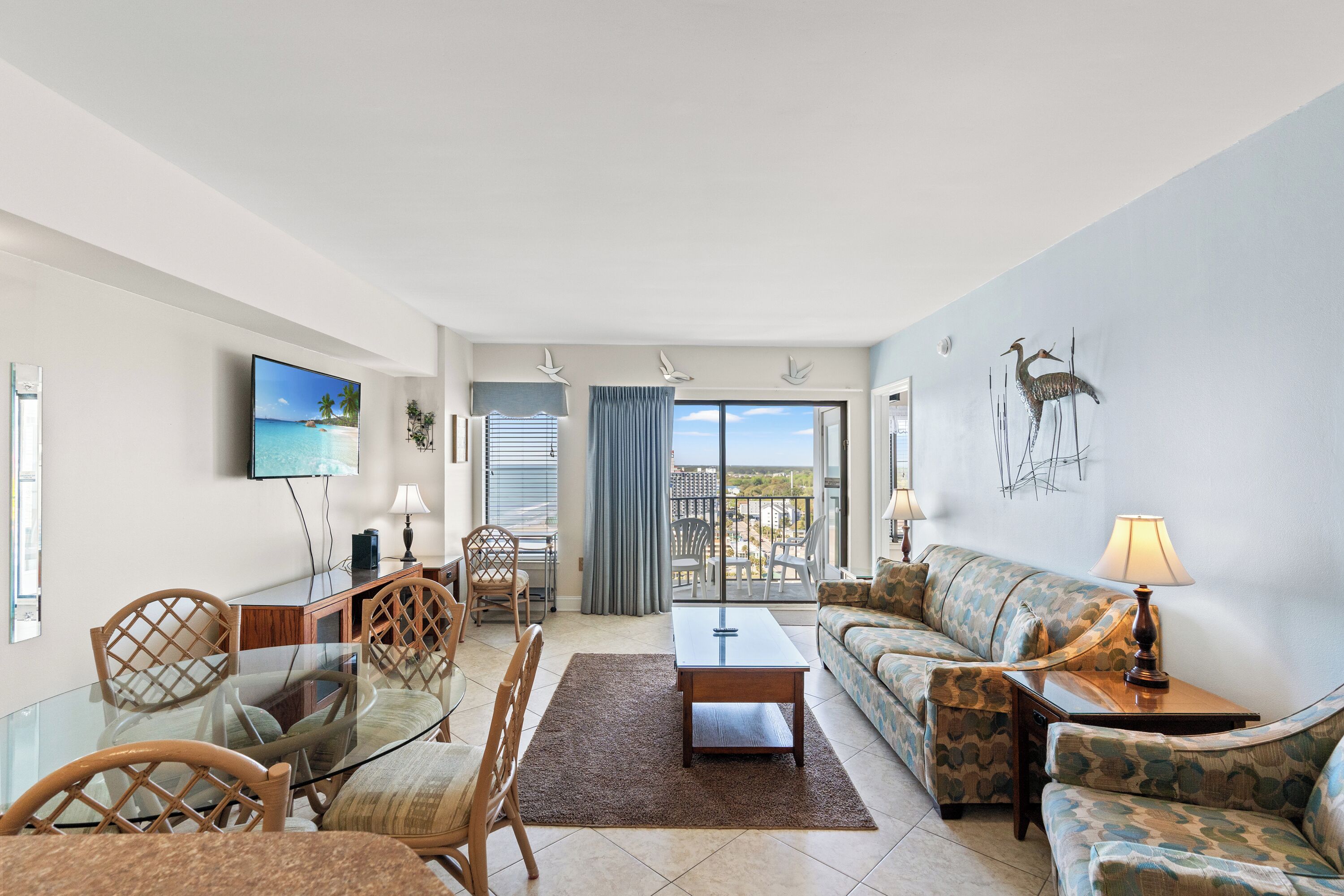 A living room with a glass table, chairs, a sofa, and a TV. There is a balcony with a view.
