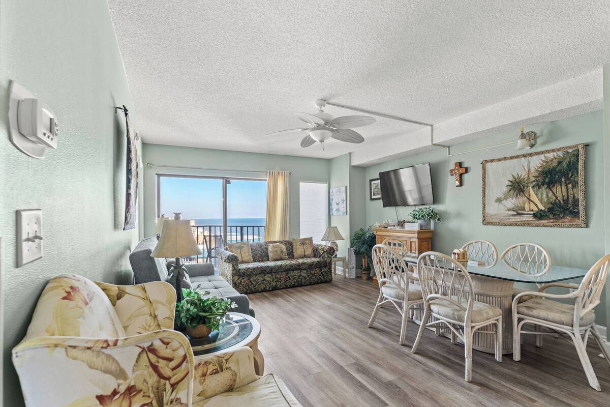 A spacious living room with a ceiling fan, a dining area with wicker chairs, a sofa, and a view of the ocean through large windows.