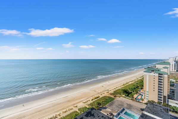 Vista de la playa con un edificio de hotel, una piscina y cielo despejado.