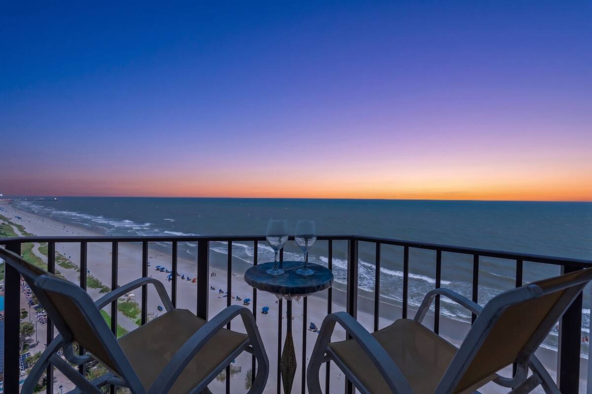 A balcony with two chairs and a small table with two wine glasses, overlooking a beach at sunset.