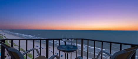 Un balcon avec deux chaises et une petite table, agrémenté de deux verres à vin, donnant sur une plage au coucher du soleil.