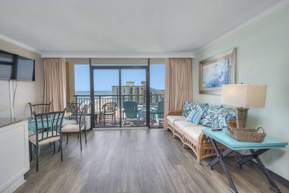 A hotel room with a balcony, a dining area with chairs and a table, a sofa with a basket, and a TV mounted on the wall.