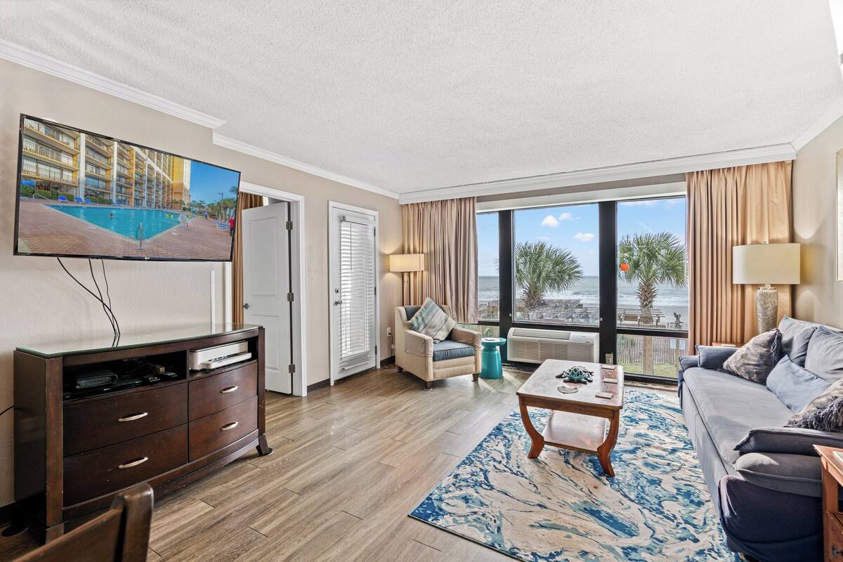 A hotel room with a television, a sofa, a coffee table, a chair, a window with a view of the beach, and a large window with curtains.