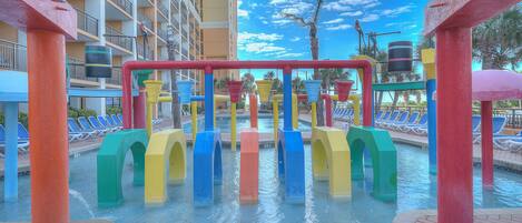 A water play area with colorful structures and a pool with lounge chairs in the background.