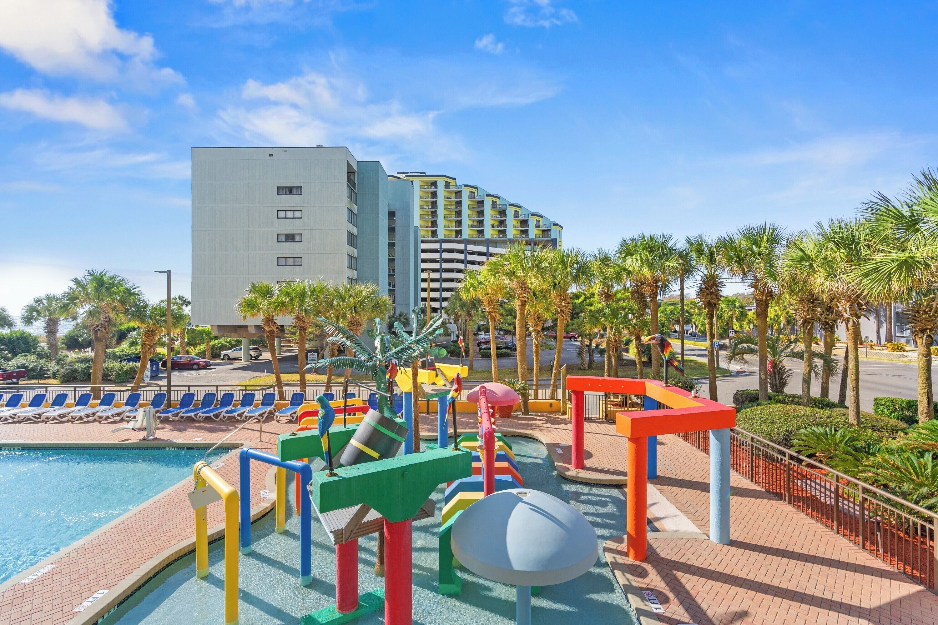 Ein Poolbereich eines Hotels mit bunten Spielgeräten, blauem Himmel und Palmen.