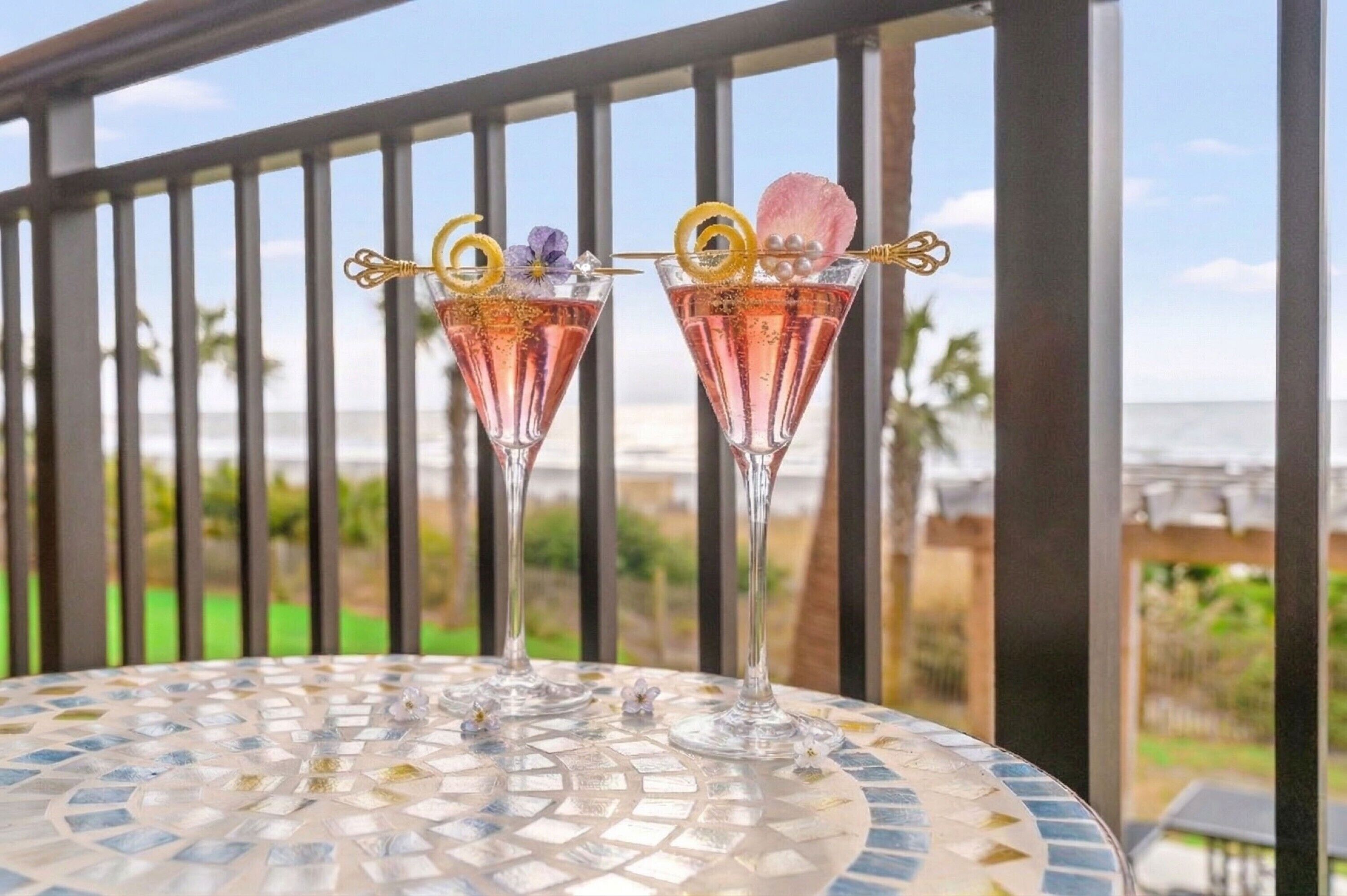 Deux verres à cocktail sont posés sur une table à motifs de mosaïque, et on peut admirer l’océan à travers la rambarde du balcon.
