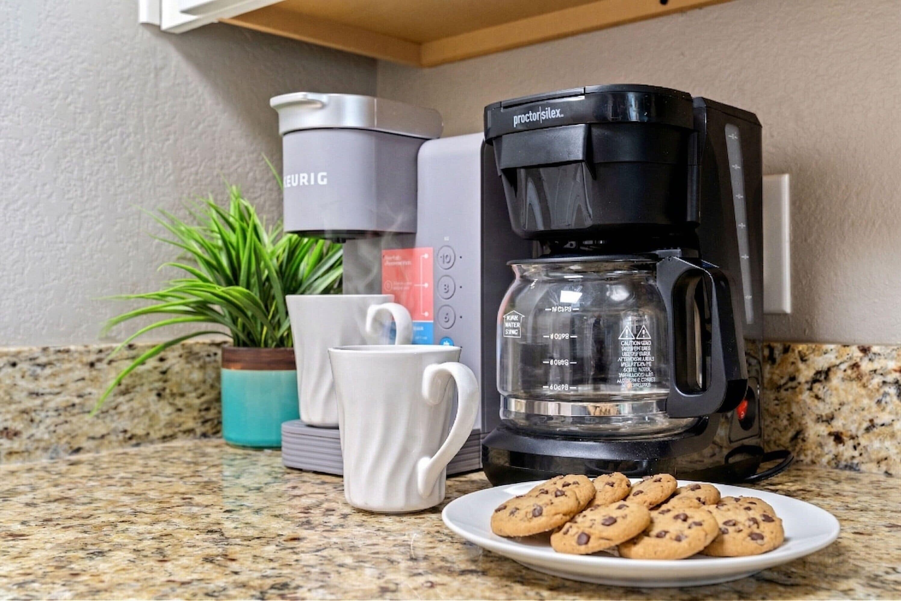 Een keuken met een koffiezetapparaat, een witte mok, een bord met koekjes en een potplant.