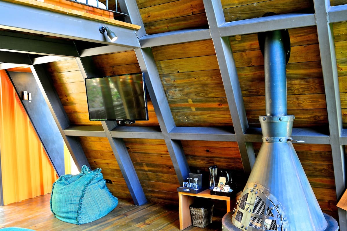 Una habitación con techo inclinado, paredes de madera, un televisor montado en la pared, una chimenea de metal y una pequeña mesa con varios objetos.