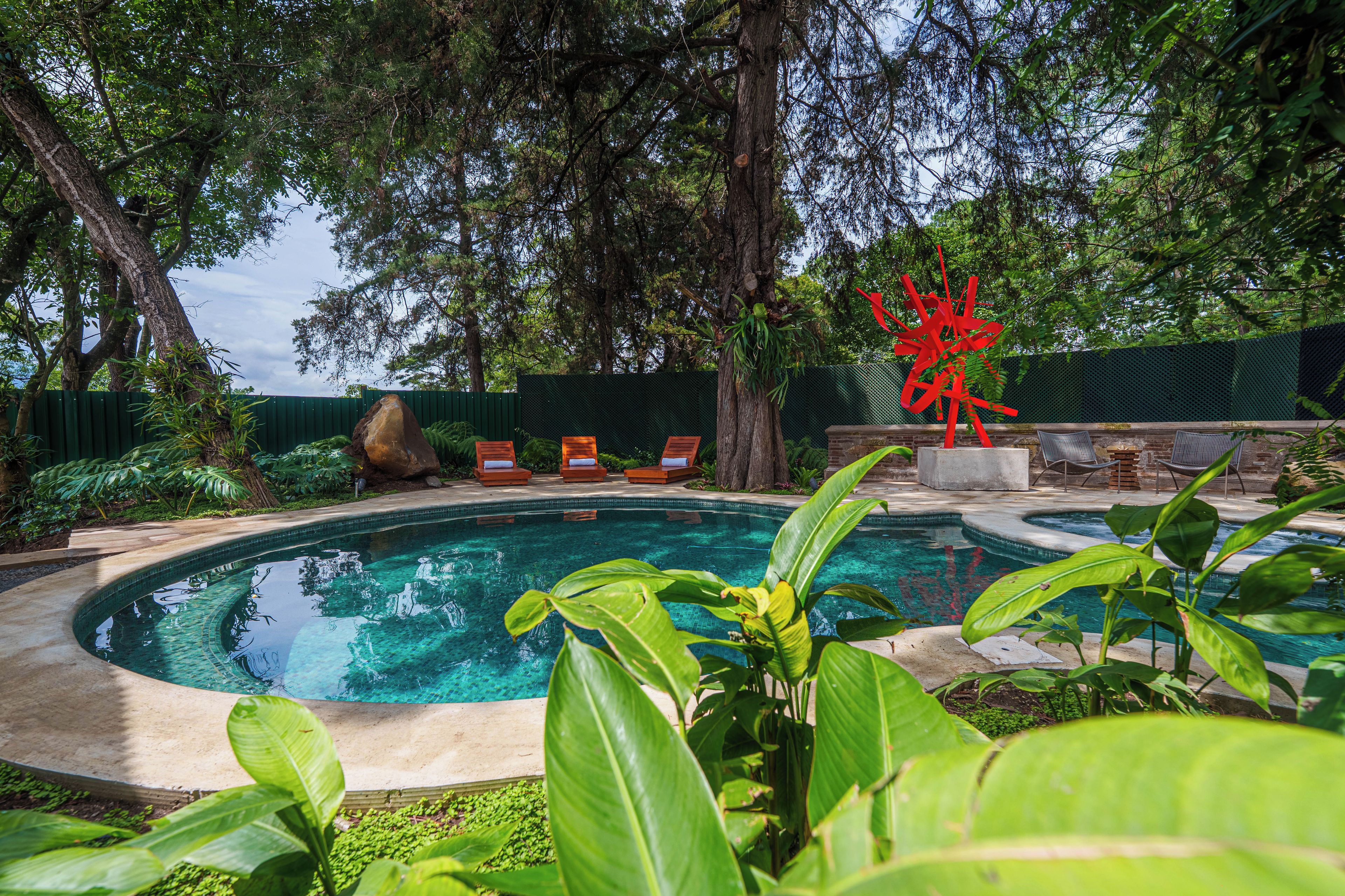 Une piscine entourée d’une végétation luxuriante et agrémentée d’une sculpture abstraite rouge.