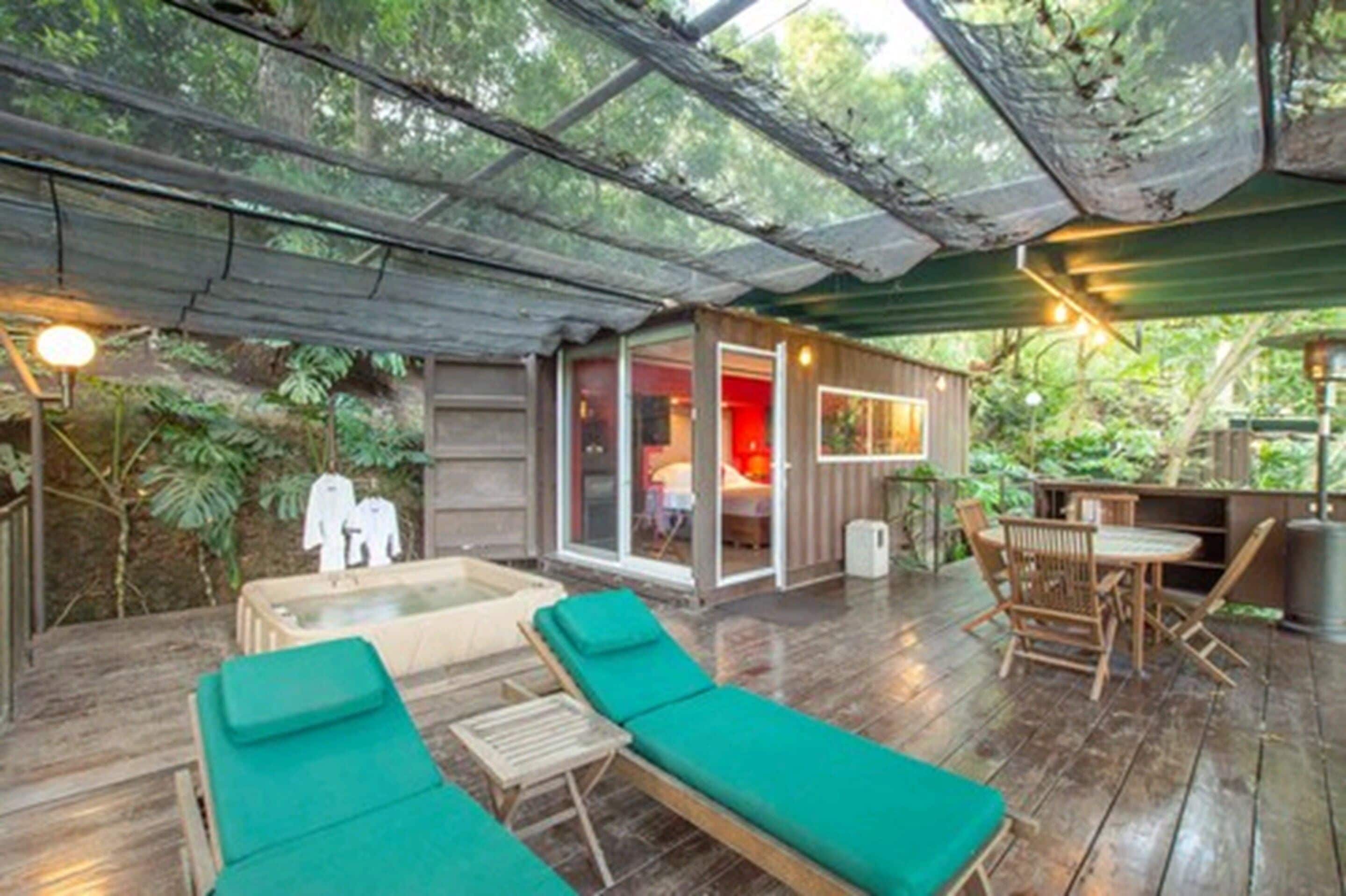 Une terrasse en bois avec des fauteuils de jardin verts, une petite table et une pièce fermée par des baies vitrées, dont l’intérieur est rouge.
