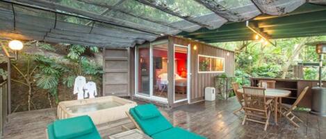 Une terrasse en bois avec des fauteuils de jardin verts, une petite table et une pièce fermée par des baies vitrées, dont l’intérieur est rouge.