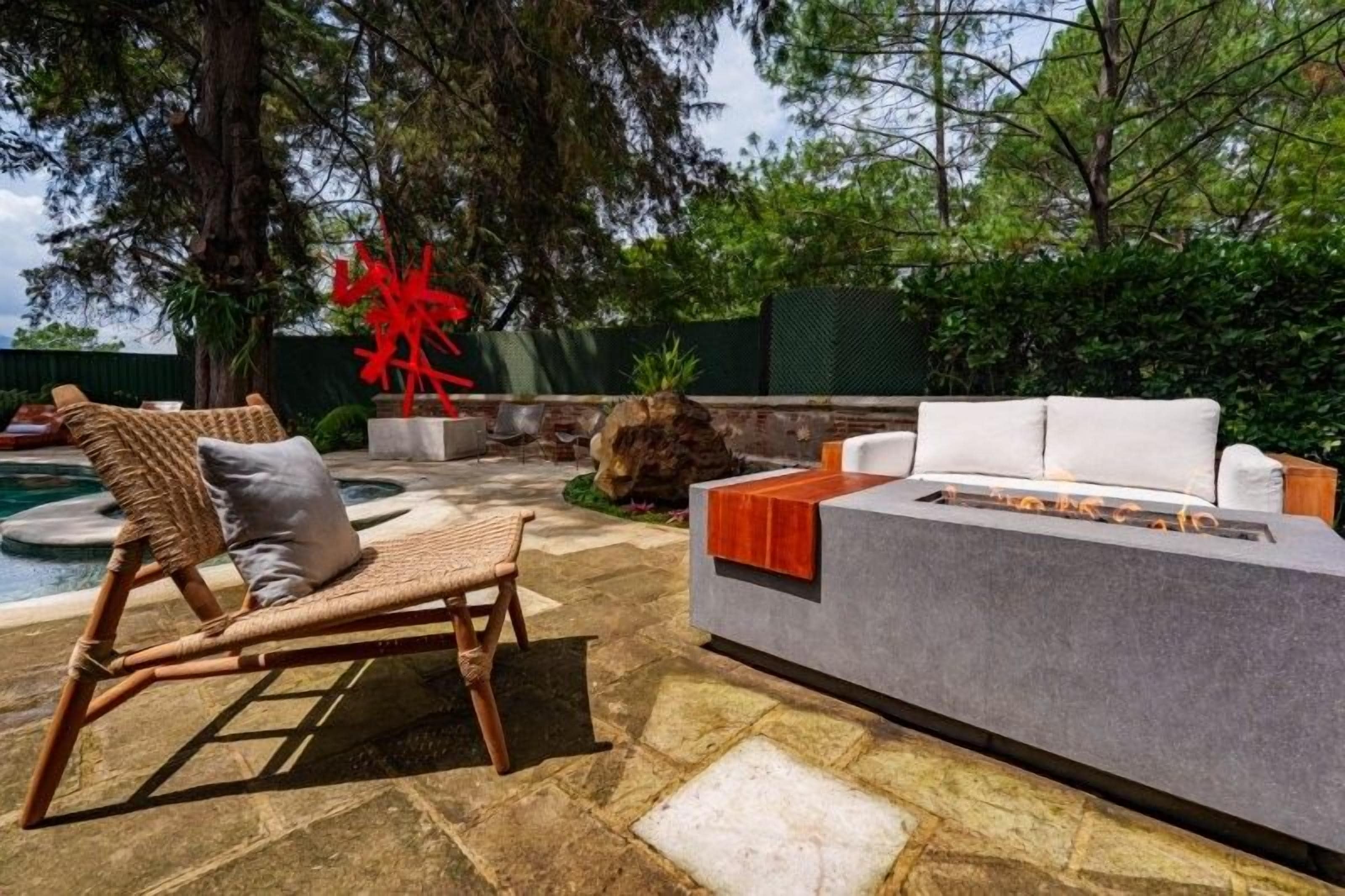 An outdoor seating area with a stone floor, a wicker lounge chair, a white sofa, and a red abstract sculpture.