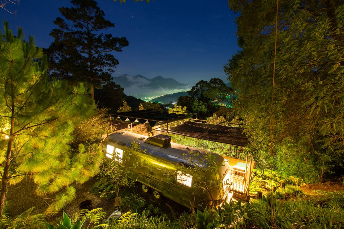 Una caravana antigua estacionada en un área boscosa, con vistas a un pueblo y montañas al fondo.