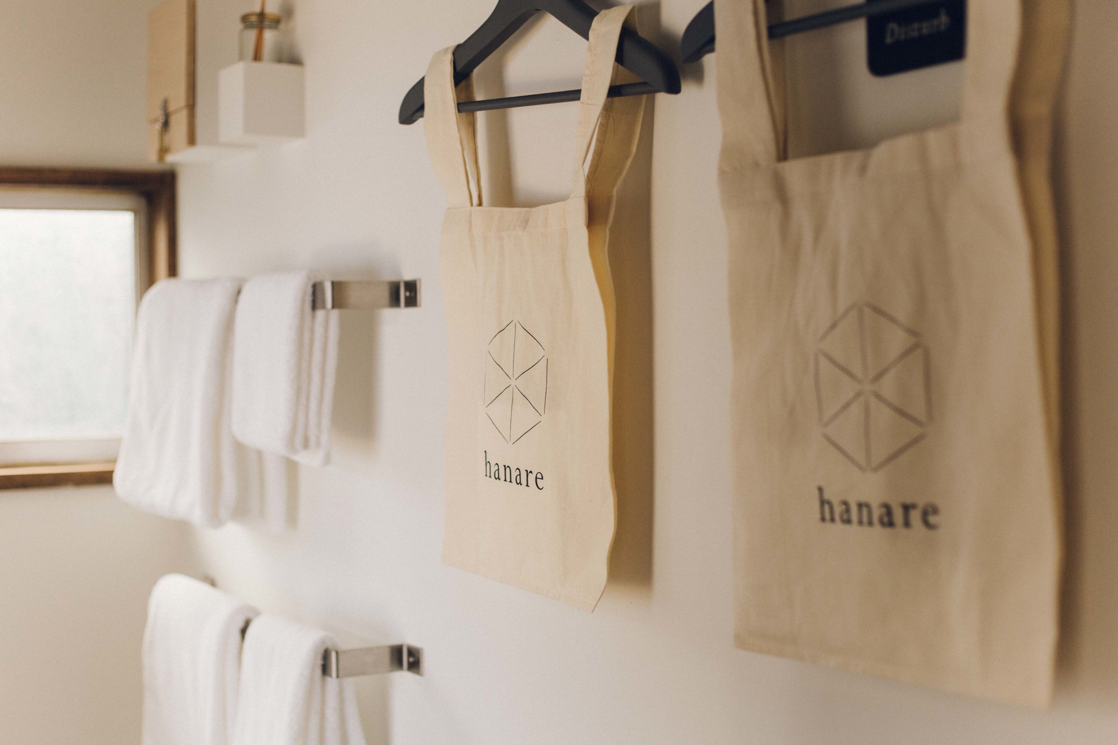 A wall with multiple hanging towels and two beige bags with the "hanare" logo.