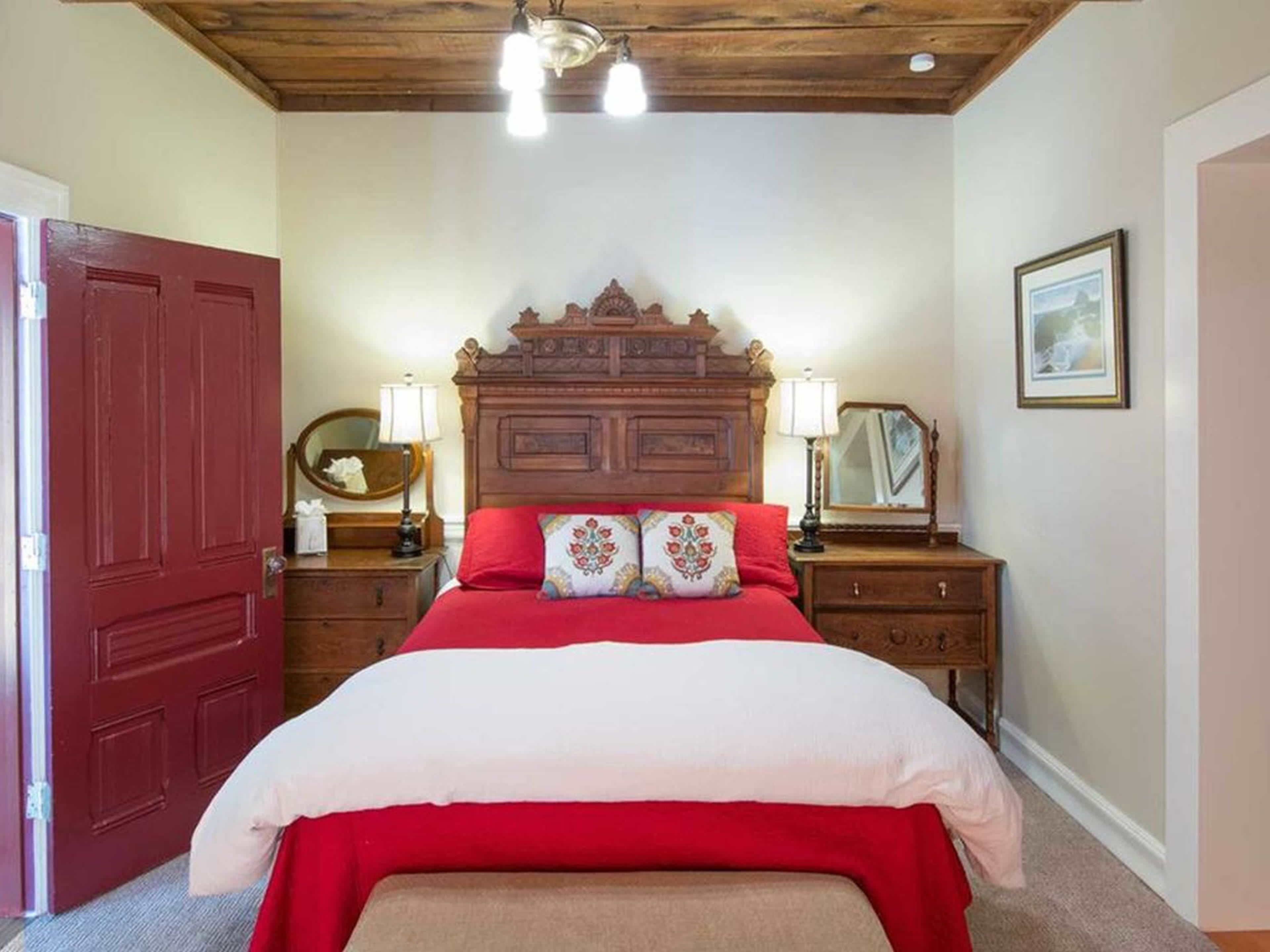 Un dormitorio con cabecera de madera, colcha roja, dos mesitas de noche con lámparas, un espejo y un cuadro enmarcado en la pared.