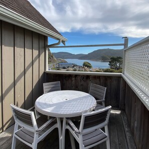 A covered outdoor patio with a round table and four chairs, overlooking a lake and hills.